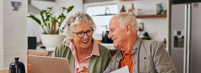 Smiling senior couple discussing bills and reviewing documents while using laptop at home